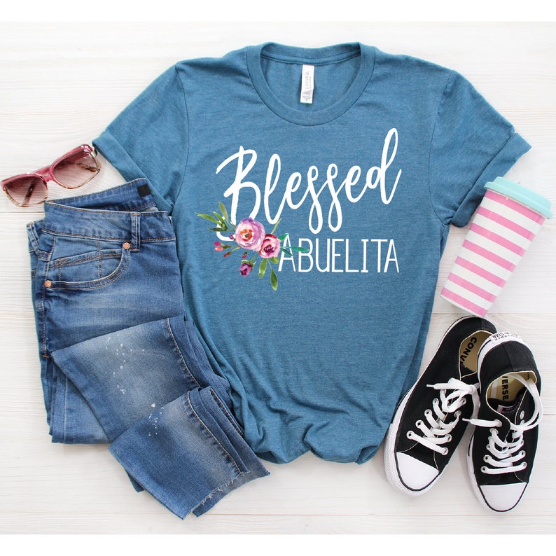 May include: A teal blue t-shirt with the text "Blessed Abuelita" in white lettering with a floral design. The shirt is laid out on a white surface with a pair of blue jeans, sunglasses, and a striped cup.
