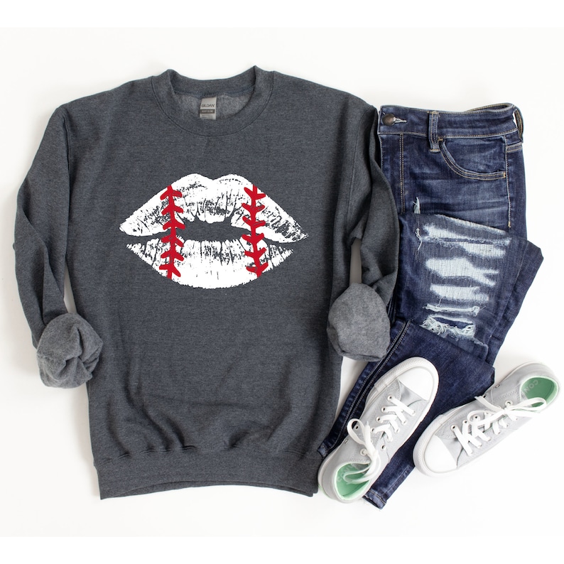 May include: A dark gray sweatshirt with a white baseball-stitched lip print. The lip is outlined in red stitching. The sweatshirt is shown with a pair of blue jeans and white sneakers.