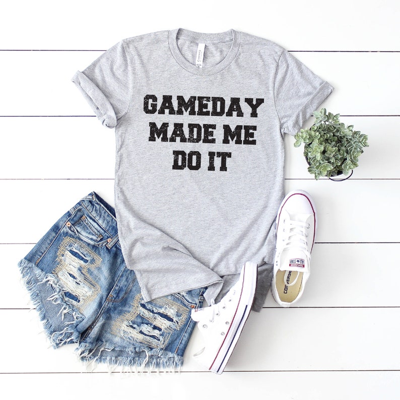 May include: A gray t-shirt with the text "GAMEDAY MADE ME DO IT" in black lettering. The shirt is paired with denim shorts and white sneakers.