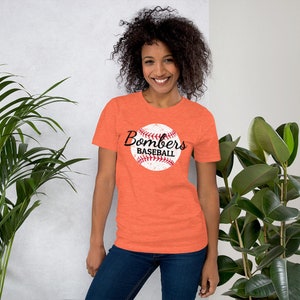 May include: A woman is wearing an orange t-shirt with a white baseball graphic and the text "Bombers Baseball".