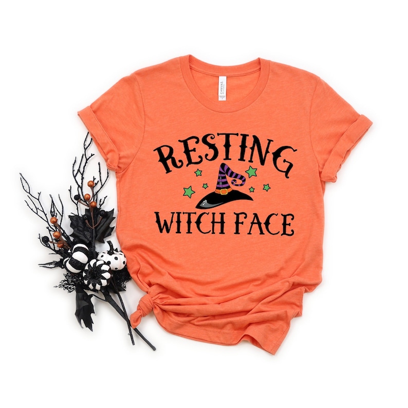 Puede incluir: Camiseta naranja con el texto "Resting Witch Face" en letras negras. Un sombrero de bruja negro con rayas moradas y blancas y estrellas verdes est&aacute; por encima del texto.