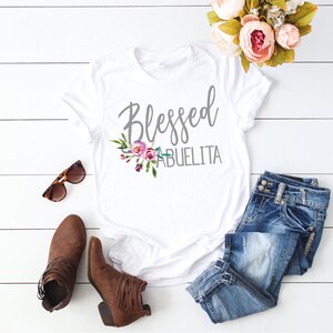 May include: White t-shirt with the text "Blessed Abuelita" in gray lettering and a floral design in pink, purple, and green.
