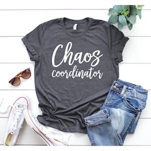 以下が含まれることがあります： 白い文字で「Chaos coordinator」と書かれた濃いグレーのTシャツ。白い木製の表面に、サングラス、ジーンズ、白いスニーカーと一緒に置かれています。