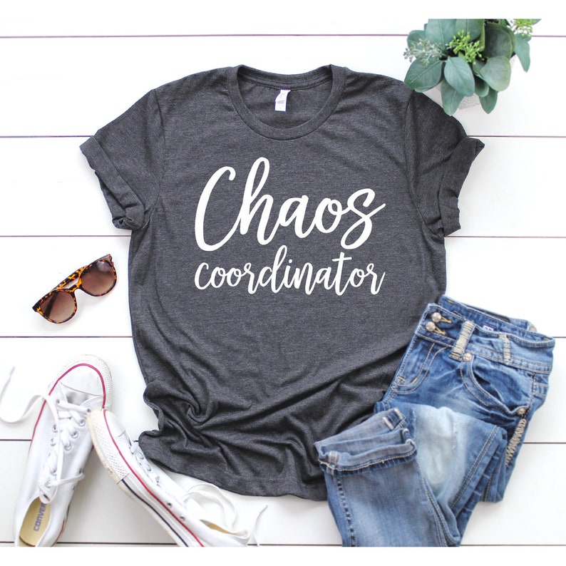 以下が含まれることがあります： 白い文字で「Chaos coordinator」と書かれた濃いグレーのTシャツ。白い木製の表面に、サングラス、ジーンズ、白いスニーカーと一緒に置かれています。