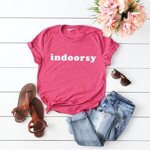 K&ouml;nnte beinhalten: Ein pinkfarbenes T-Shirt mit dem Wort "indoorsy" in wei&szlig; auf der Vorderseite. Das Shirt liegt auf einer wei&szlig;en Holzoberfl&auml;che mit einem Paar braunen Sandalen, einer blauen Jeans und einer Sonnenbrille.
