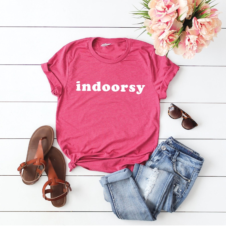 K&ouml;nnte beinhalten: Ein pinkfarbenes T-Shirt mit dem Wort "indoorsy" in wei&szlig; auf der Vorderseite. Das Shirt liegt auf einer wei&szlig;en Holzoberfl&auml;che mit einem Paar braunen Sandalen, einer blauen Jeans und einer Sonnenbrille.