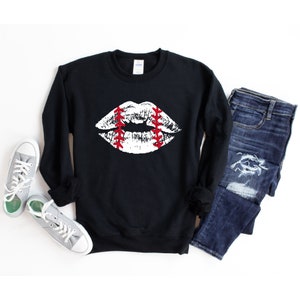 May include: Black sweatshirt with a white baseball-themed lips graphic. The sweatshirt is laid flat on a white surface with a pair of grey sneakers and blue jeans.