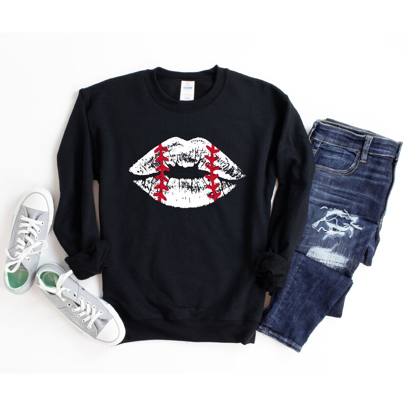 May include: Black sweatshirt with a white baseball-themed lips graphic. The sweatshirt is laid flat on a white surface with a pair of grey sneakers and blue jeans.