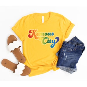 Pu&ograve; includere: Una maglietta gialla con una grafica arcobaleno che dice "Kansas City" in un font retr&ograve;.