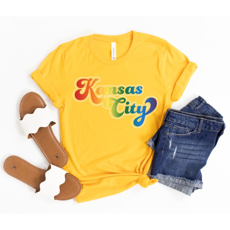 Pu&ograve; includere: Una maglietta gialla con una grafica arcobaleno che dice "Kansas City" in un font retr&ograve;.