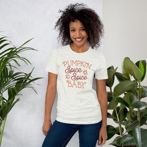 Op de afbeelding: Een lichtgrijs t-shirt met de tekst "PUMPKIN Spice Spice BABY" in bruin script. Het shirt heeft korte mouwen en een ronde hals. Het model draagt een blauwe spijkerbroek.