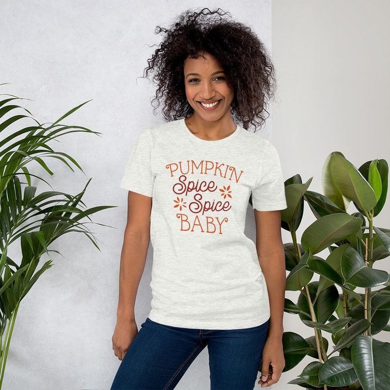Op de afbeelding: Een lichtgrijs t-shirt met de tekst "PUMPKIN Spice Spice BABY" in bruin script. Het shirt heeft korte mouwen en een ronde hals. Het model draagt een blauwe spijkerbroek.