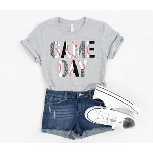 May include: A gray t-shirt with the words "GAME DAY" printed on it. The letters are made up of a baseball pattern with red stitching and black and white leopard print.