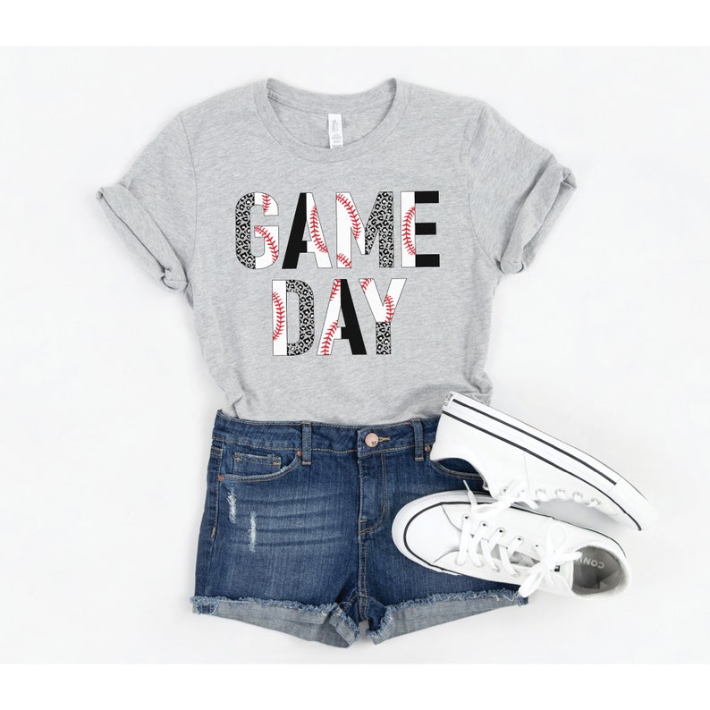 May include: A gray t-shirt with the words "GAME DAY" printed on it. The letters are made up of a baseball pattern with red stitching and black and white leopard print.