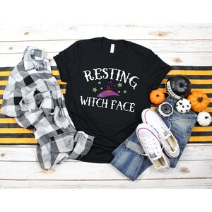 Puede incluir: Camiseta negra con el texto "Resting Witch Face" en blanco. Un sombrero de bruja morado con estrellas verdes est&aacute; por encima del texto. La camiseta est&aacute; colocada sobre una superficie de madera blanca con una camisa de cuadros, pantalones vaqueros y calabazas.