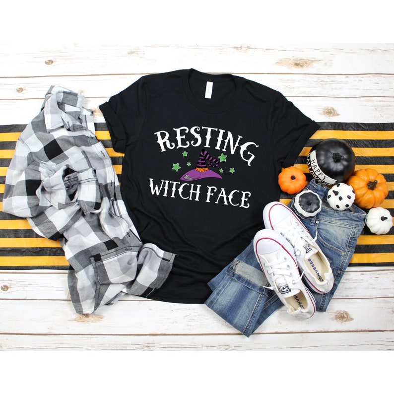 Puede incluir: Camiseta negra con el texto "Resting Witch Face" en blanco. Un sombrero de bruja morado con estrellas verdes est&aacute; por encima del texto. La camiseta est&aacute; colocada sobre una superficie de madera blanca con una camisa de cuadros, pantalones vaqueros y calabazas.