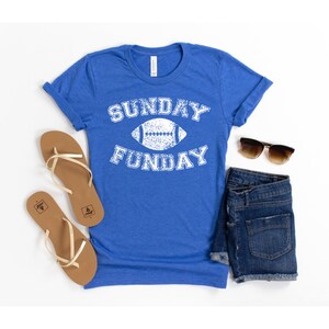 Op de afbeelding: Een koninklijk blauw t-shirt met een witte, verweerde afbeelding met de tekst "SUNDAY FUNDAY" met een voetbal in het midden.