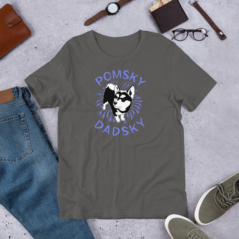 May include: A dark gray t-shirt with a white cartoon Pomsky dog wearing a blue collar. The dog is surrounded by blue fireworks. The text "Pomsky Dadsky" is written in blue letters above and below the dog.