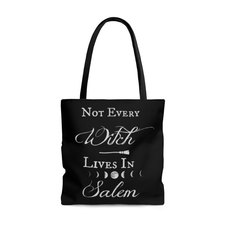 May include: Black tote bag with white text that reads "Not Every Witch Lives In Salem". The text is in a cursive font and there is a small broom graphic above the word "Witch". There are also four moon phases in a row below the word "Salem".