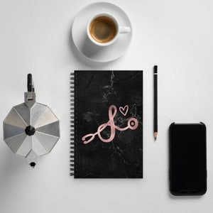 May include: A black spiral notebook with a marble pattern and a rose gold stethoscope and heart design. The notebook is on a white surface with a coffee cup, a pen, and a black phone.