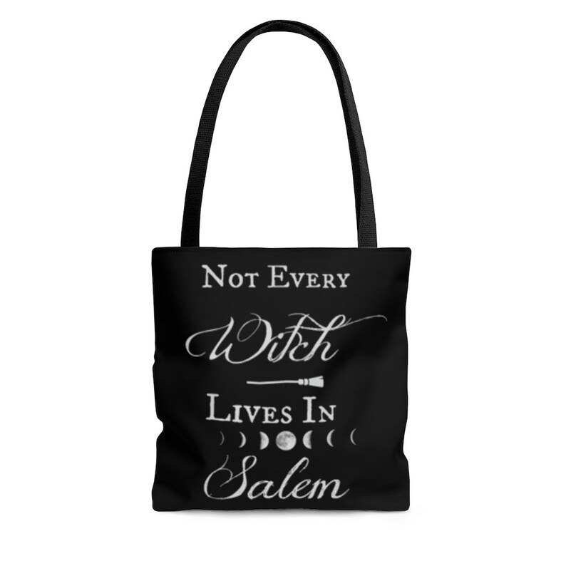 May include: Black tote bag with white text that reads "Not Every Witch Lives In Salem". The text is in a cursive font and includes a small broom graphic. There are also four white crescent moons below the text.