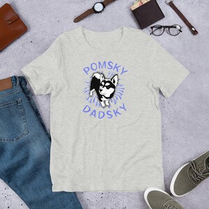 May include: A heather grey t-shirt with a black and white cartoon Pomsky dog wearing a blue collar. The dog is surrounded by blue fireworks. The text "Pomsky Dadsky" is written in blue letters around the dog.