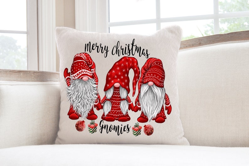 Puede incluir: Almohada blanca con un dise&ntilde;o de gnomo navide&ntilde;o rojo y blanco. La almohada presenta tres gnomos con sombreros rojos y barbas blancas. El texto "merry christmas gnomies" est&aacute; impreso en la almohada.