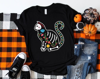 Könnte beinhalten: Schwarzes T-Shirt mit einem farbenfrohen Totenkopf-Katzen-Design. Die Katze ist weiß umrandet und hat ein Skelett-Design mit floralen Akzenten. Das Shirt liegt auf einer schwarz-weißen karierten Oberfläche mit einem Paar schwarzer Sneakers und einer blauen Jeans.