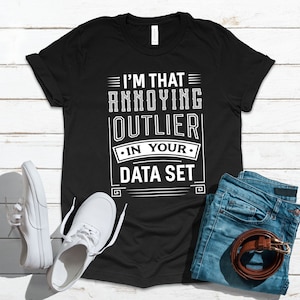 Puede incluir: Camiseta negra con texto blanco que dice "Soy ese outlier molesto en tu conjunto de datos". La camiseta está colocada sobre una superficie de madera blanca con un par de zapatillas blancas y unos vaqueros azules con un cinturón marrón.
