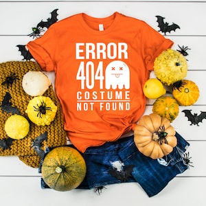 Puede incluir: Camiseta naranja con un gráfico blanco que dice "Error 404 Disfraz no encontrado" con una ilustración de fantasma. La camiseta está rodeada de calabazas, calabazas y murciélagos sobre un fondo de madera blanca.