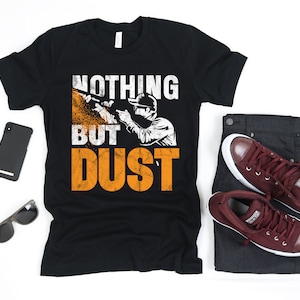May include: Black t-shirt with a distressed white graphic of a person using a power tool with the text "Nothing But Dust".