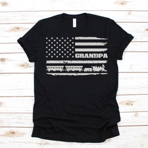 May include: Black t-shirt featuring a distressed American flag design with the word "GRANDPA" printed in white. A train is depicted in front of the flag.