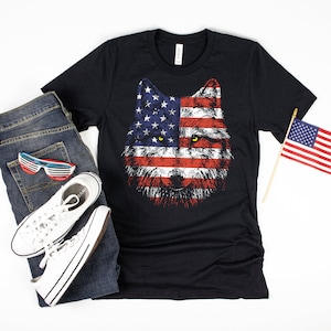 Puede incluir: Una camiseta negra con un gráfico de la cara de un lobo. La cara del lobo está hecha con la bandera estadounidense. La camiseta se combina con jeans azules, zapatillas blancas y una pequeña bandera estadounidense.