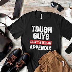 Puede incluir: Camiseta negra con texto blanco que dice "Tough Guys Don't Need An Appendix". La camiseta está colocada sobre una superficie de madera con pantalones marrones, zapatos negros, una bolsa de cuero marrón, gafas de sol y un llavero.
