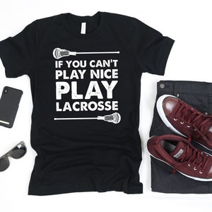 May include: Black t-shirt with the text "IF YOU CAN'T PLAY NICE PLAY LACROSSE" in white. The shirt is paired with dark grey jeans and maroon trainers. A black smartphone and sunglasses are also in the image.