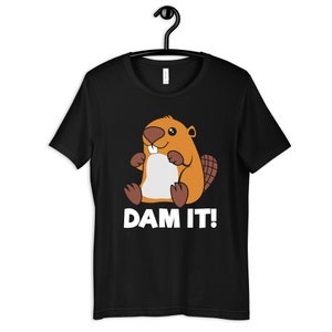 May include: Black t-shirt featuring a cartoon beaver wearing a brown and orange fur coat. The beaver is sitting with its paws crossed and has a toothy grin. The text "DAM IT!" is printed in white below the beaver.