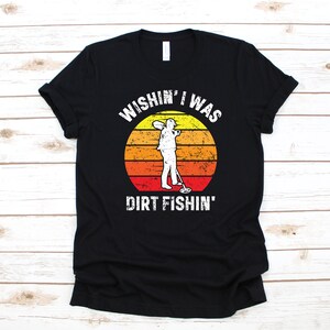 以下が含まれることがあります： 「Wishin' I Was Dirt Fishin'」というテキストと、金属探知機を持った人物のヴィンテージスタイルのグラフィックが描かれた黒いTシャツ。