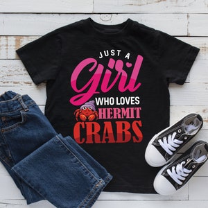May include: Black t-shirt with pink text that reads "Just a Girl Who Loves Hermit Crabs". A cartoon hermit crab is pictured above the text. The shirt is on a white wooden surface with a pair of blue jeans and black sneakers.