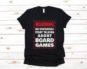 May include: Black t-shirt with a red and white warning sign graphic that says "Warning May Spontaneously Start Talking About Board Games".