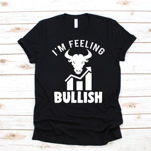 May include: Black t-shirt with a white graphic. The graphic features a bull's head above the words "I'M FEELING" and "BULLISH", with an upward trending graph. The shirt is displayed on a light wood grain surface.