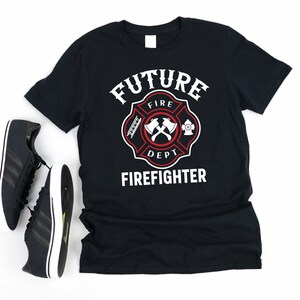 Può includere: Maglietta nera con una grafica bianca di un emblema di pompiere. L'emblema presenta una croce rossa con asce, una scala e un idrante. Il testo "FUTURE FIRE DEPT. FIREFIGHTER" è stampato sotto l'emblema.