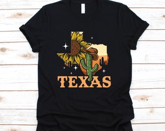 Puede incluir: Camiseta negra con un gráfico del estado de Texas, un girasol, un cactus con un sombrero de vaquero y la palabra "TEXAS" en naranja.