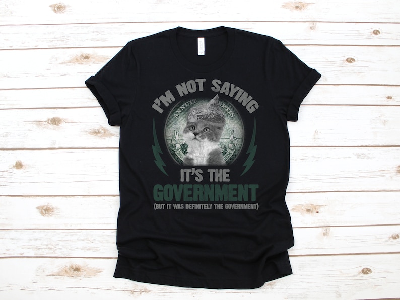 May include: Black t-shirt with a green and white graphic of a cat wearing a dollar bill as a hat. The text on the shirt reads "I'm not saying it's the government (but it was definitely the government)"