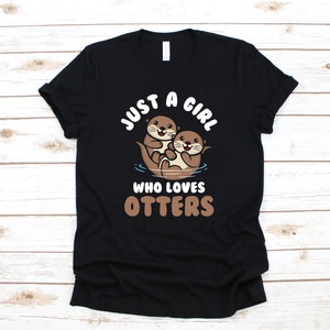 Op de afbeelding: Zwart T-shirt met een witte afbeelding van twee otters en de tekst "Just a girl who loves otters".