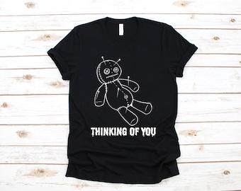 May include: Black t-shirt featuring a white illustration of a voodoo doll with pins. The text "THINKING OF YOU" is printed below the doll in a distressed font. The shirt is made of a soft material.