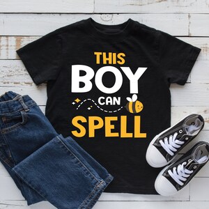 Puede incluir: Camiseta negra con texto amarillo que dice "This Boy Can Spell" y una abeja de dibujos animados. La camiseta está sobre una superficie de madera blanca con un par de pantalones vaqueros azules y zapatillas negras.