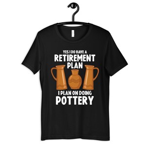 以下が含まれることがあります： 黒いTシャツに白い文字で「はい、私は退職プランを持っています。陶芸をするつもりです」と書かれています。テキストは3つの茶色の陶器の壺で囲まれています。