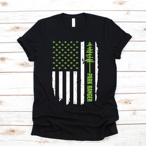 May include: Black t-shirt with a distressed American flag design in white and green. The flag has a green pine tree and the words "Park Ranger" written vertically on the right side.