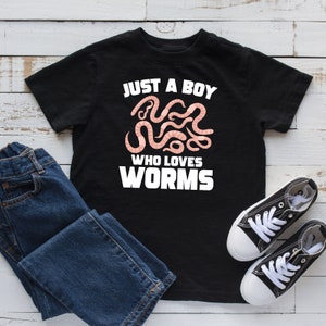 Puede incluir: Camiseta negra con texto blanco que dice "Just a boy who loves worms". El texto está rodeado por un grupo de gusanos rosas.