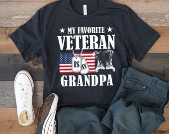 May include: Navy blue t-shirt with the text "MY FAVORITE VETERAN IS MY GRANDPA" in white. The design features an American flag, dog tags, and military boots. The image also includes white trainers and blue jeans.